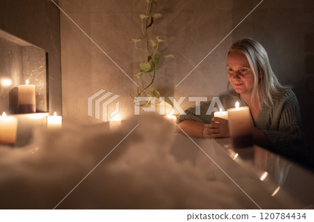 A serene woman relaxes in a softly lit bathroom, surrounded by glowing candles. She gazes thoughtfully at the candlelight, creating a warm and tranquil atmosphere 120784434