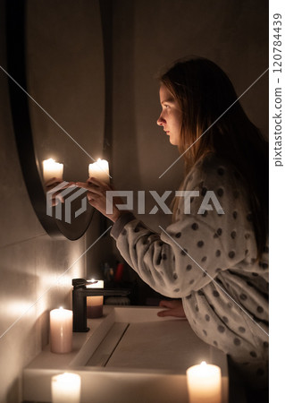 A young woman in soft lighting holds a candle near a mirror, her finger touching her reflection. The warm glow of the candles creates a calm and intimate atmosphere. 120784439