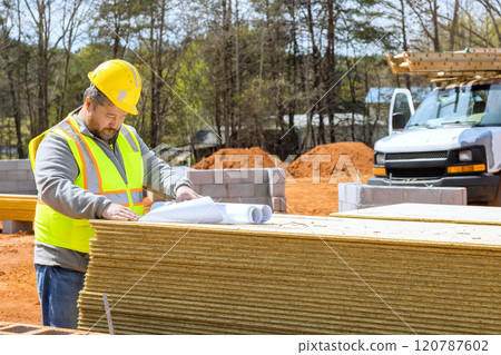 Construction supervisor worker in safety vest, hard hat studies blueprints on stack of materials at residential building site heavy machinery Construction supervisor worker in safety vest, hard hat studies blueprints on stack of materials at residential building site heavy machinery 120787602
