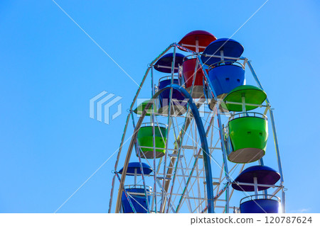 Colorful Ferris wheel with red, blue, green gondolas against bright blue sky. 120787624