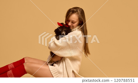 Portrait of beautiful young woman wearing light cozy winter sweater and red socks, hugging her adorable purebred black Poodle dog against studio background 120788551