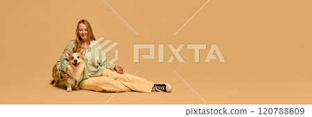 Portrait of happy young woman in mint color cardigan posing with her adorable smiling Corgi dog in matching light green jacket again studio background 120788609