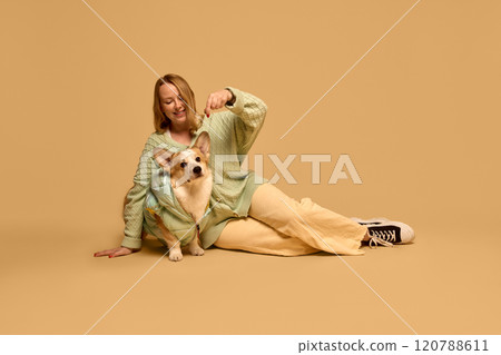 Beautiful young woman wearing cozy light green cardigan, posing, playing with her adorable Corgi dog wearing matching green jacket against studio background Beautiful young woman wearing cozy light green cardigan, posing, playing with her adorable Corgi dog wearing matching green jacket against studio background 120788611