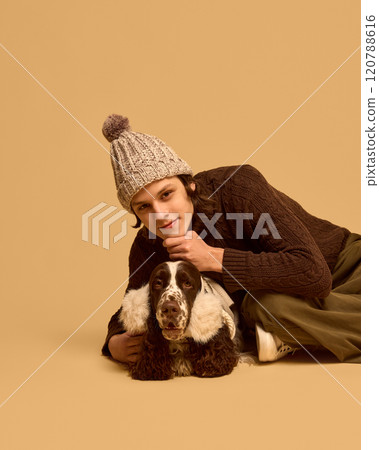 Portrait of young man in warm, cozy chocolate sweater posing with his adorable calm dog wearing warm headwear, against beige studio background 120788616