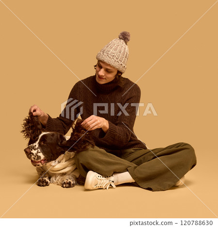 Young man in cozy winter clothes, chocolate sweater and knitted hat, playing with his purebred dog wearing sweater, against studio background Young man in cozy winter clothes, chocolate sweater and knitted hat, playing with his purebred dog wearing sweater, against studio background 120788630