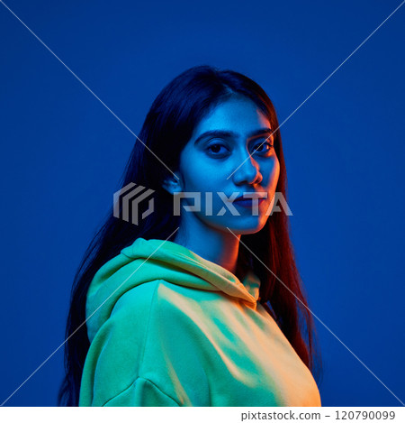 Portrait of young Indian woman wearing yellow hoodie, posing with calm expression against blue studio background in neon light 120790099