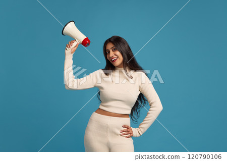 Young smiling Indian woman in light clothes, holding megaphone with smile against blue studio background. News, announcement Young smiling Indian woman in light clothes, holding megaphone with smile against blue studio background. News, announcement 120790106