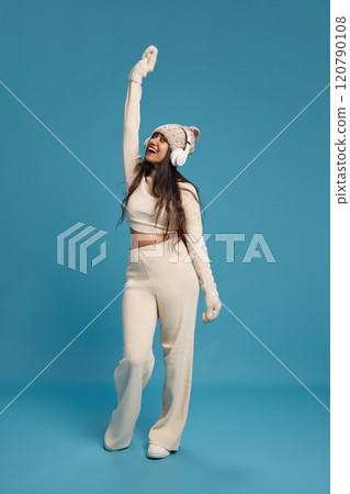 Full-length portrait of happy young Indian in knitted hat, listening to music in headphones and dancing against blue studio background Full-length portrait of happy young Indian in knitted hat, listening to music in headphones and dancing against blue studio background 120790108