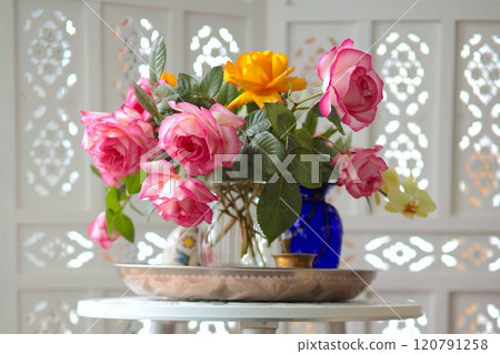 Pink and yellow roses in glass vase on metal tray on white round table. Background white wooden folding screen room divider. Floral arrangement as decoration for interior Pink and yellow roses in glass vase on metal tray on white round table. Background white wooden folding screen room divider. Floral arrangement as decoration for interior 120791258