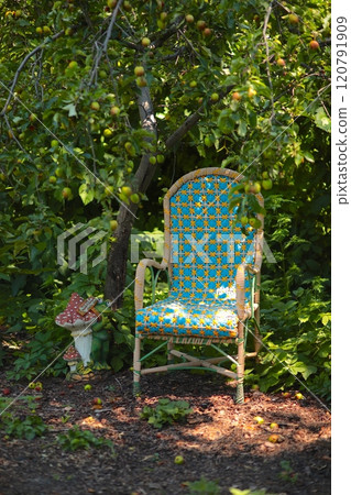 Blue wicker chair located under apple tree in garden next to mushroom garden figure and against backdrop of green foliage that add peace. Carefree morning in garden. Retro chair in garden 120791909