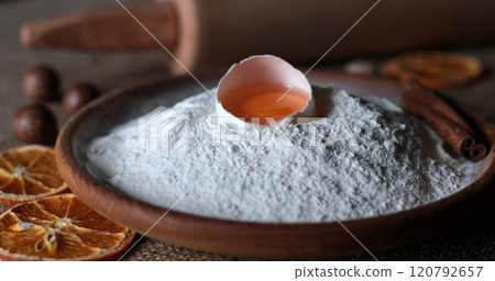 Rustic style still life of organic food ingredients for homemade baking with white flour and egg yolk inside half of broken egg shell. Flour powder pile in traditional wooden bowl. Dried orange slices 120792657