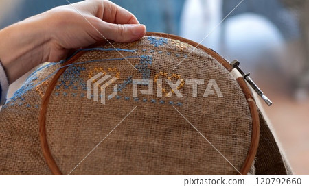 Female hand hold wooden embroidery hoop with cross stitch pattern on rough burlap fabric. Yellow blue modern folk motif embroidery with yellow hearts. Traditional Ukrainian needlework on canvas Female hand hold wooden embroidery hoop with cross stitch pattern on rough burlap fabric. Yellow blue modern folk motif embroidery with yellow hearts. Traditional Ukrainian needlework on canvas 120792660