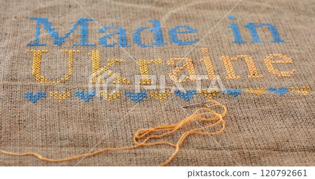 Handmade burlap fabric with blue yellow cross stitch embroidery text Made in Ukraine. Selective focus embroidery with heart pattern in Ukrainian colors on burlap canvas. Needle with yellow thread Handmade burlap fabric with blue yellow cross stitch embroidery text Made in Ukraine. Selective focus embroidery with heart pattern in Ukrainian colors on burlap canvas. Needle with yellow thread 120792661