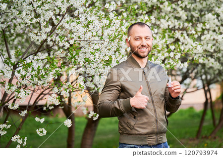 Man allergic using medical eyes drops, suffering from seasonal allergy at spring in blossoming garden. Portrait of smiling man showing thumbs up in front of blooming tree. Spring allergy concept. Man allergic using medical eyes drops, suffering from seasonal allergy at spring in blossoming garden. Portrait of smiling man showing thumbs up in front of blooming tree. Spring allergy concept. 120793974