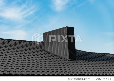 A modern, black chimney sits atop a black, tiled roof. The chimney is rectangular with vertical lines. The background is a blue sky with white clouds 120794574