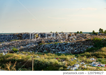 Bulldozers maneuver through a substantial landfill, compacting trash under a clear afternoon sky, surrounded by sparse vegetation and distant trees Bulldozers maneuver through a substantial landfill, compacting trash under a clear afternoon sky, surrounded by sparse vegetation and distant trees 120794627