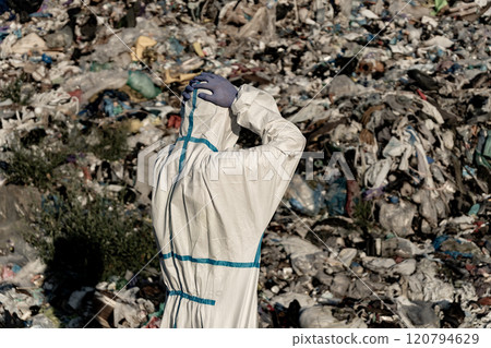 A worker wearing a protective suit examines a sprawling landfill overflowing with waste materials under bright daylight, highlighting the urgent issue of pollution and waste management 120794629