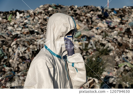 A person dressed in protective clothing and gloves examines a vast area filled with waste. The worker stands amidst piles of garbage, highlighting environmental concerns 120794630