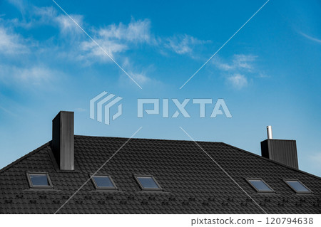 A modern house with a black tiled roof showcases several skylights against a bright blue sky. The daylight highlights the architecture and roofing design 120794638