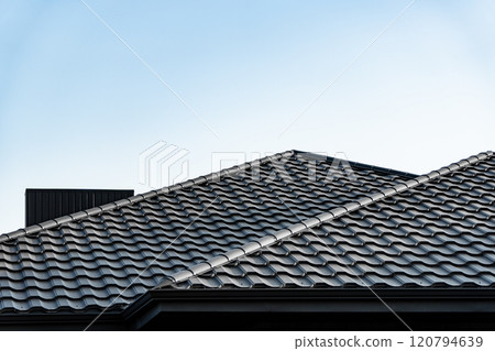 Two intersecting rooftops featuring textured tiles rise under a bright blue sky, emphasizing modern construction and design aesthetics in residential architecture Two intersecting rooftops featuring textured tiles rise under a bright blue sky, emphasizing modern construction and design aesthetics in residential architecture 120794639