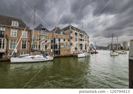 Serene harbor with moored boats under a dramatic cloudy sky. Quaint historic buildings and trees line the waterfront, reflecting in the calm green water, creating peaceful scene. Dordrecht landscape. 120794813