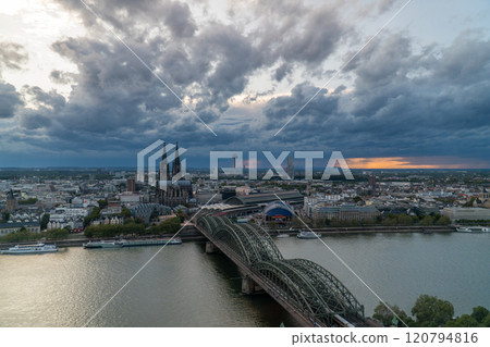 A scenic view of Cologne, Germany, featuring the iconic Cologne Cathedral, the Hohenzollern Bridge, the Rhine River, and a dramatic cloudy sky with hints of sunset light on the horizon. 120794816