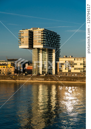 Modern riverside building with a unique architectural design reflects on the calm water under a clear blue sky. The warm sunlight highlights its structure and nearby surroundings. Landscape of Cologne Modern riverside building with a unique architectural design reflects on the calm water under a clear blue sky. The warm sunlight highlights its structure and nearby surroundings. Landscape of Cologne 120794817