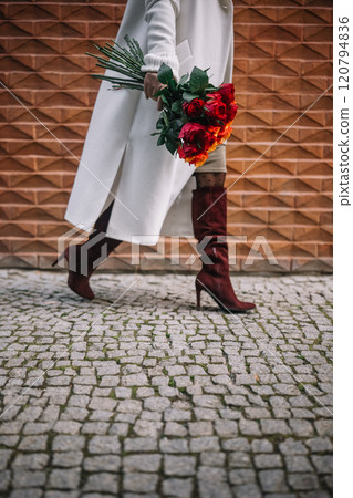 Woman walks on cobblestone path while carrying vibrant flower bouquet in elegant boots 120794836