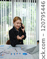 A woman is talking on her cell phone while sitting at a desk with a laptop 120795446