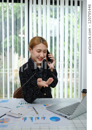 A woman is talking on her cell phone while sitting at a desk with a laptop A woman is talking on her cell phone while sitting at a desk with a laptop 120795446