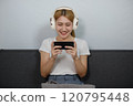 A woman is sitting on a couch and playing a game on her phone 120795448