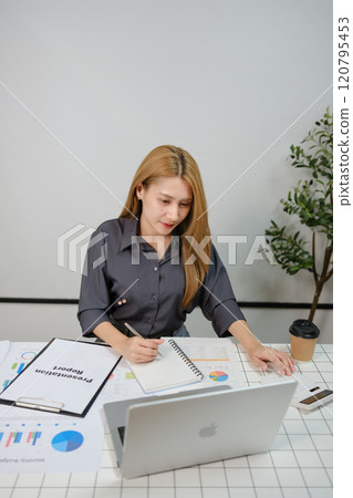 A woman is sitting at a desk with a laptop and a notebook A woman is sitting at a desk with a laptop and a notebook 120795453