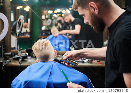 Hairdresser shaving boy's head. Competent barber using electric shaver at barbershop. Little client getting stylish men's haircut. 120795548