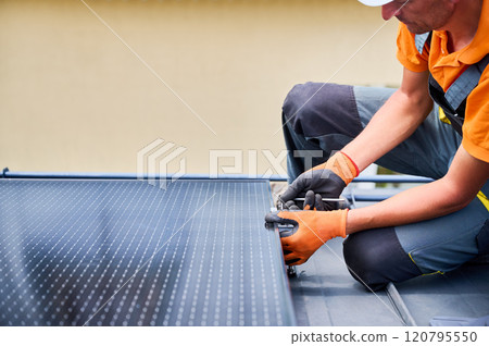 Worker building photovoltaic solar panel system on rooftop of house. Man engineer in gloves installing solar module with help of hex key outdoors. Alternative and renewable energy generation concept. 120795550