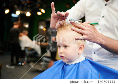 Man barber cutting little boy's hair using comb and scissors. Child getting haircut from adult male, likely barber. Professional hairdresser and cute client at modern barbershop. Man barber cutting little boy's hair using comb and scissors. Child getting haircut from adult male, likely barber. Professional hairdresser and cute client at modern barbershop. 120795553