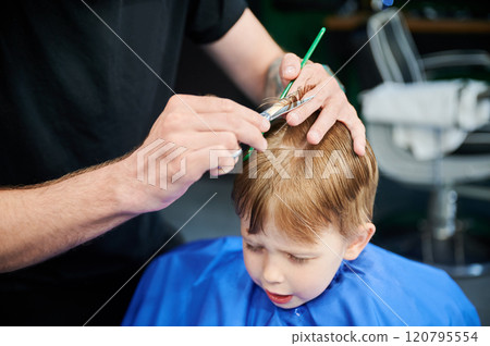 Man barber cutting little boy's hair using comb and scissors. Child getting haircut from adult male, likely barber. Professional hairdresser and cute client at modern barbershop. 120795554