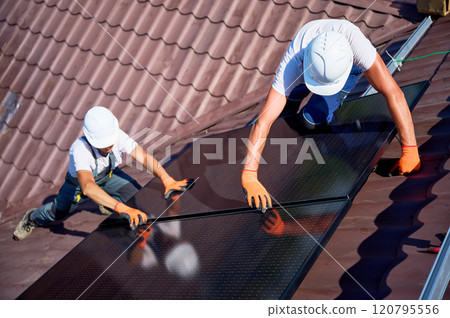 Workers building solar panel system on rooftop of house. Two men installers in helmets installing photovoltaic solar module outdoors. Alternative, green and renewable energy generation concept. 120795556