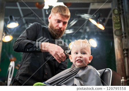 Cute little boy getting haircut. Blond curious kid first time at barbershop. Professional hairdresser making stylish haircut by shaver. 120795558