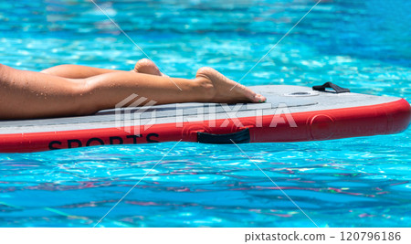 Close-up of legs on a paddleboard in a tropical pool. Concept of aquatic relaxation and fitness Close-up of legs on a paddleboard in a tropical pool. Concept of aquatic relaxation and fitness 120796186