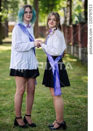 Two female graduates embrace while wearing sashes on their last day of school, celebrating their academic success in a park 120797013