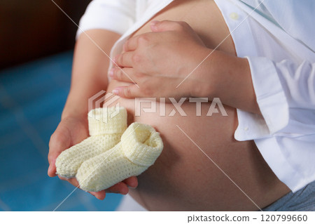 A pregnant woman gently cradles her baby bump while holding pair of soft, knitted booties, symbolizing joy and anticipation of new life 120799660