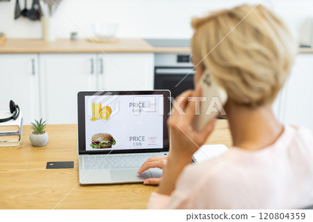 Caucasian woman using phone to order food online while browsing delivery website on laptop. She sits at table in modern kitchen, exploring menu options and discounts. Caucasian woman using phone to order food online while browsing delivery website on laptop. She sits at table in modern kitchen, exploring menu options and discounts. 120804359
