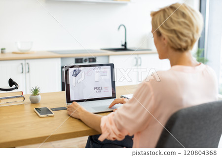 Caucasian woman in her 30s shopping online for clothes using laptop in modern kitchen. Focused on laptop screen displaying clothing prices, she is seated at wooden table with smartphone and notepad. 120804368