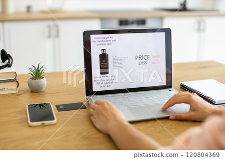 Caucasian woman using laptop to order cosmetics online, showcasing convenience of internet shopping. Modern workspace features laptop, smartphone, and minimalistic decor. 120804369