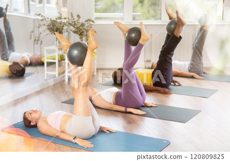 Students lying on floor, holding fitball in raised straight legs, rolling fitball in shins and feet 120809825