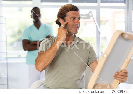 Satisfied male client examines face after rejuvenating procedure with African-American cosmetologist Satisfied male client examines face after rejuvenating procedure with African-American cosmetologist 120809893