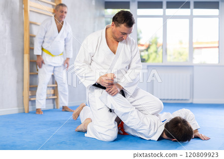 Two men practice grabbing and throwing sports mats during judo training under the guidance of an experienced mature coach 120809923