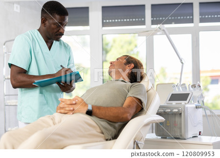 African male doctor conducts survey of man patient after rejuvenating procedure 120809953