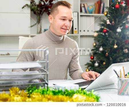 Positive man office worker using laptop during Christmastime Positive man office worker using laptop during Christmastime 120810016