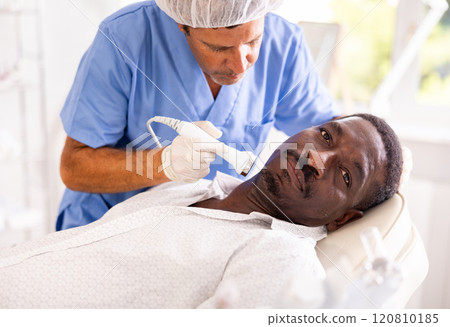 Male specialist uses ultrasonic import nozzle to care African man client face skin 120810185